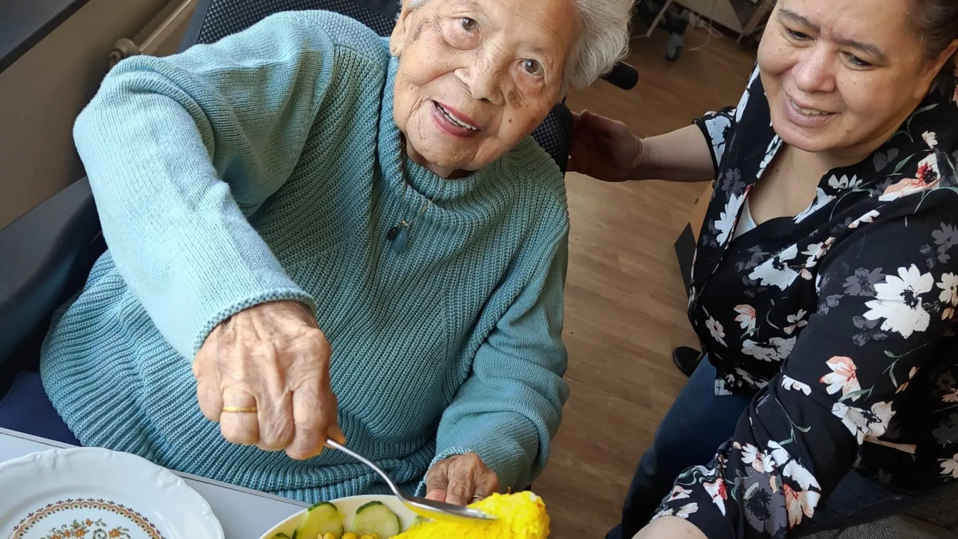 Een oudere vrouw snijdt een traditionele gele rijsttaart, bijgestaan door een andere vrouw.